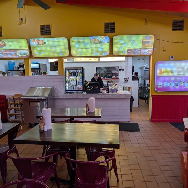 Interior of a casual restaurant with yellow walls, red trim, menu boards, and empty tables with chairs.