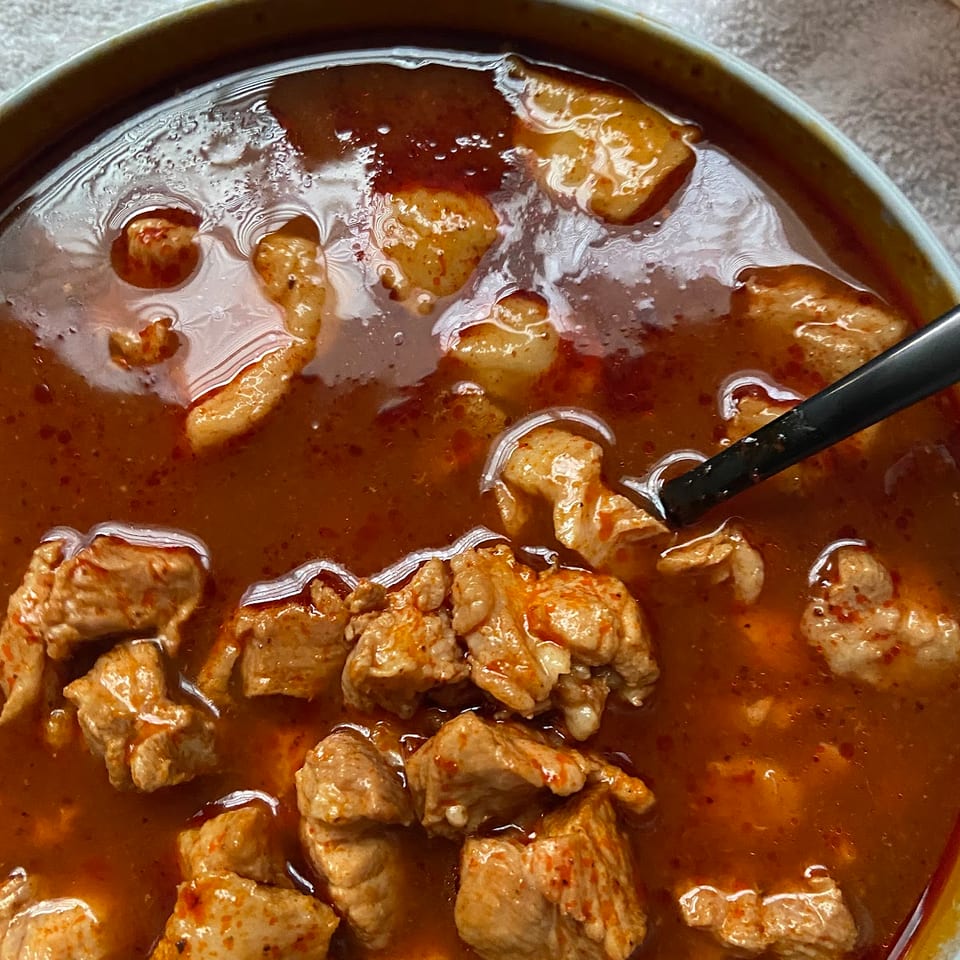 Close-up of a bowl of red stew with chunks of meat and a black spoon.