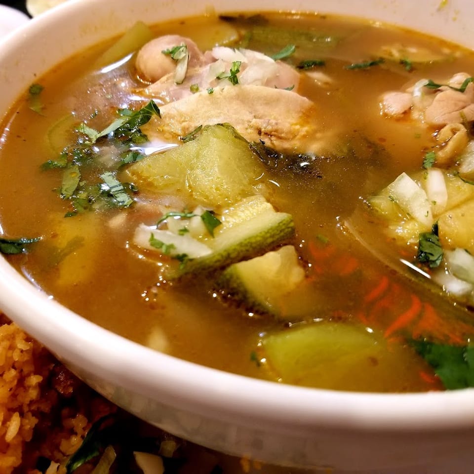 Close-up of a bowl of chicken soup with zucchini, onions, and cilantro, next to rice.