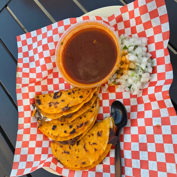 Birria tacos with consommé and diced onions on red and white checkered paper.