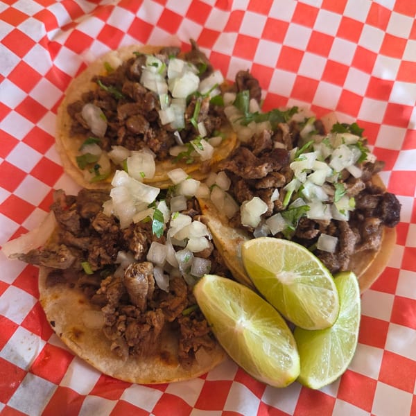Three street tacos with meat, diced onions, cilantro, and lime wedges on red and white checkered paper.