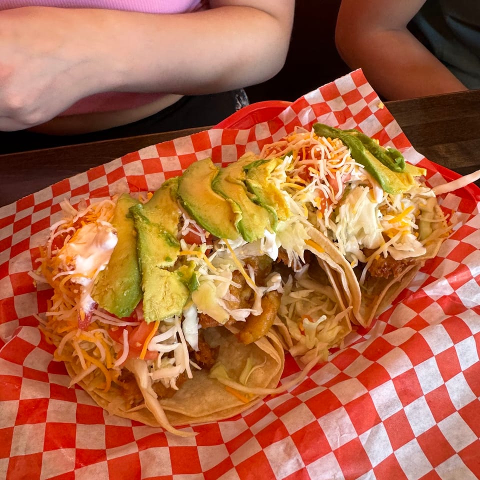 Three loaded tacos with avocado, shredded cheese, and cabbage in a red and white checkered basket.
